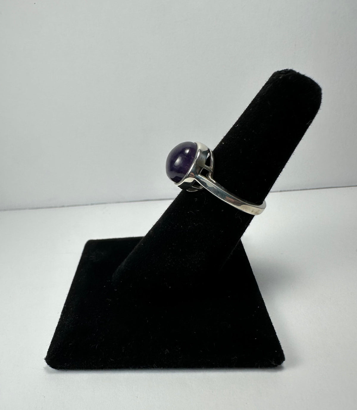 Silver ring with a round, polished deep purple amethyst stone set in a simple bezel, displayed upright on a black velvet finger stand against a white background.