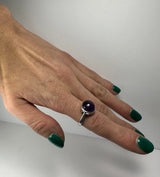Hand with green nail polish wearing a silver ring set with a round polished purple amethyst stone on the middle finger.