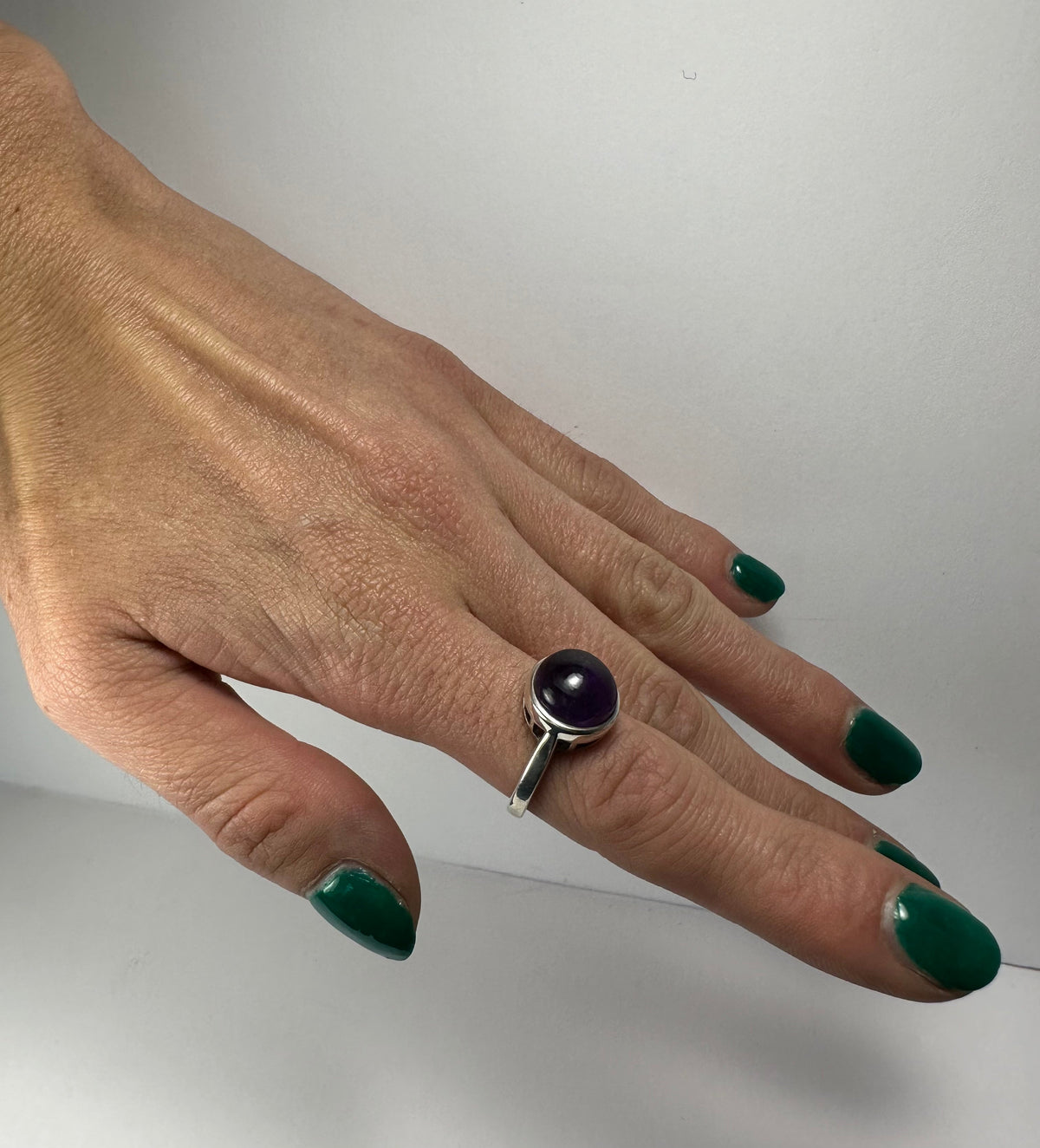 Hand with green nail polish wearing a silver ring set with a round polished purple amethyst stone on the middle finger.