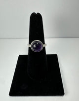 Silver ring with a round, polished dark purple amethyst stone set in a bezel, displayed upright on a black velvet stand against a white background.