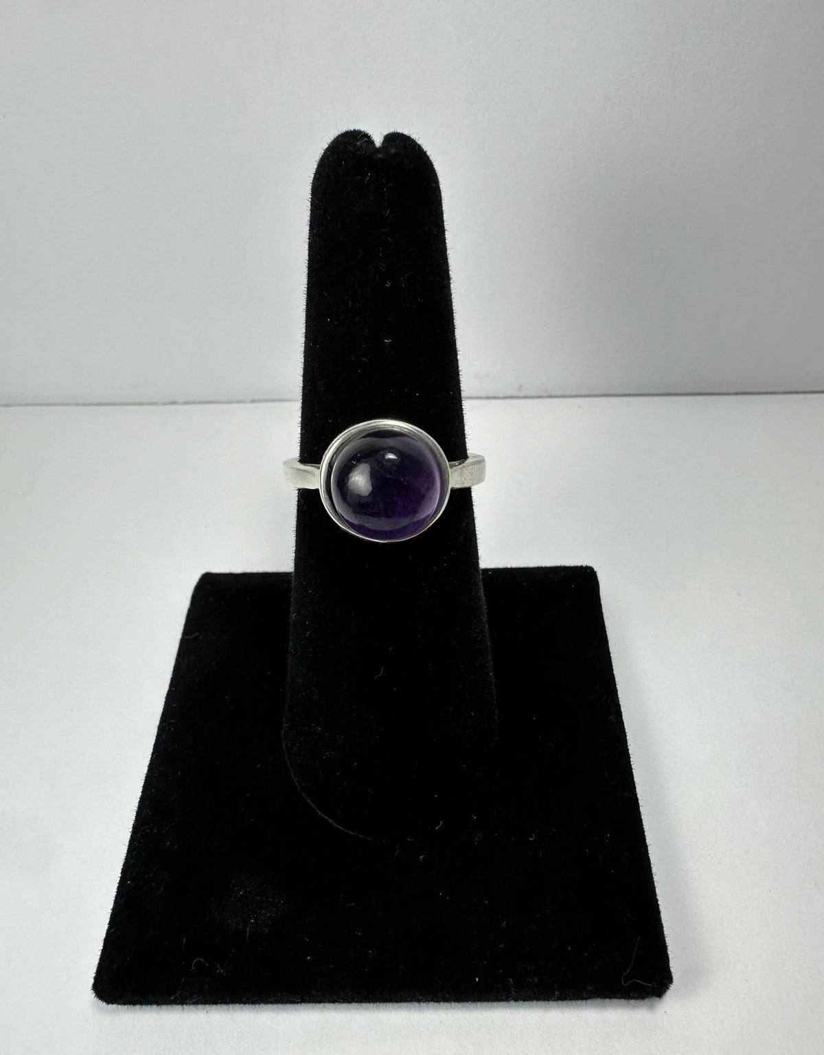 Silver ring with a round, polished dark purple amethyst stone set in a bezel, displayed upright on a black velvet stand against a white background.