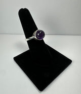 Silver ring with a round, polished purple amethyst stone displayed on a black velvet stand against a plain white background.