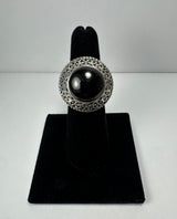 Round garnet ring with a glossy deep red stone set in an ornate silver band featuring intricate filigree patterns, displayed on a black velvet ring stand.