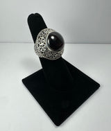 Silver ring with an ornate filigree band and a polished, round deep red garnet stone set in a domed bezel, displayed on a black velvet ring stand.