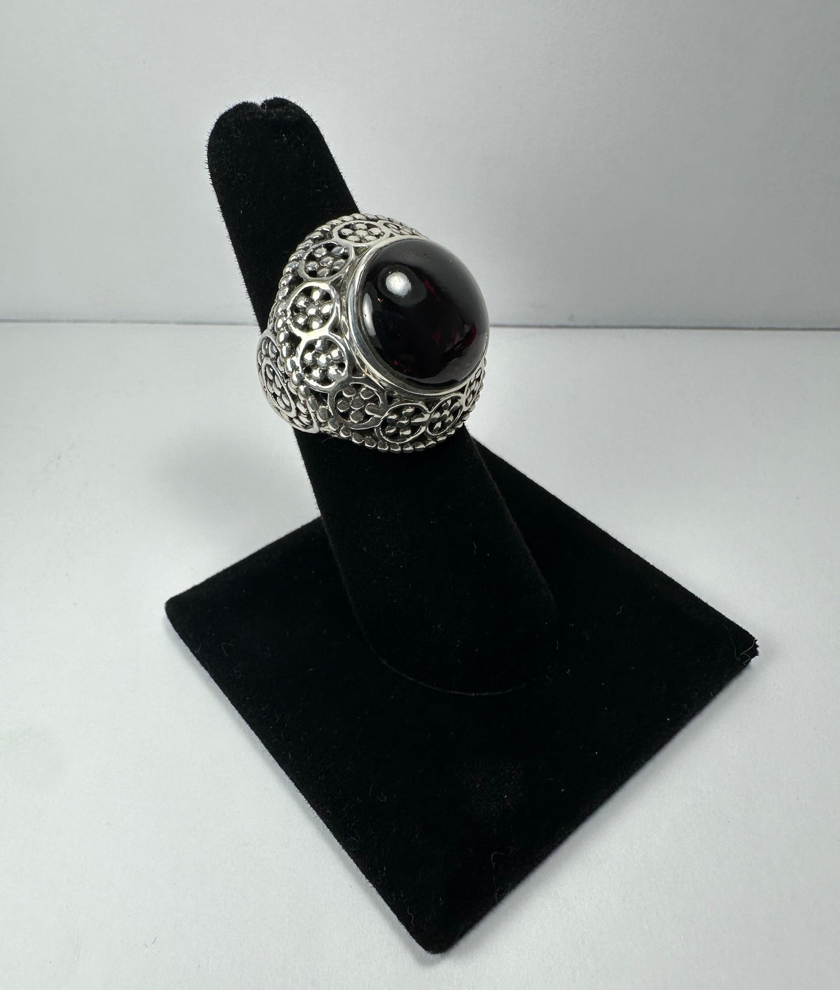 Silver ring with an ornate filigree band and a polished, round deep red garnet stone set in a domed bezel, displayed on a black velvet ring stand.