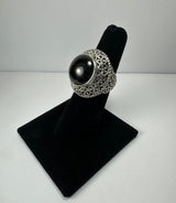 Silver ring with intricate filigree detailing and a large, glossy black cabochon gemstone, displayed on a black velvet stand against a plain white background.