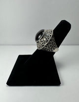 Silver ring with intricate circular filigree patterns and a smooth black garnet cabochon center, displayed on a black angled ring stand.