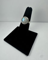 Silver ring with a round iridescent moonstone set in a smooth bezel, displayed upright on a black velvet stand against a plain white background.