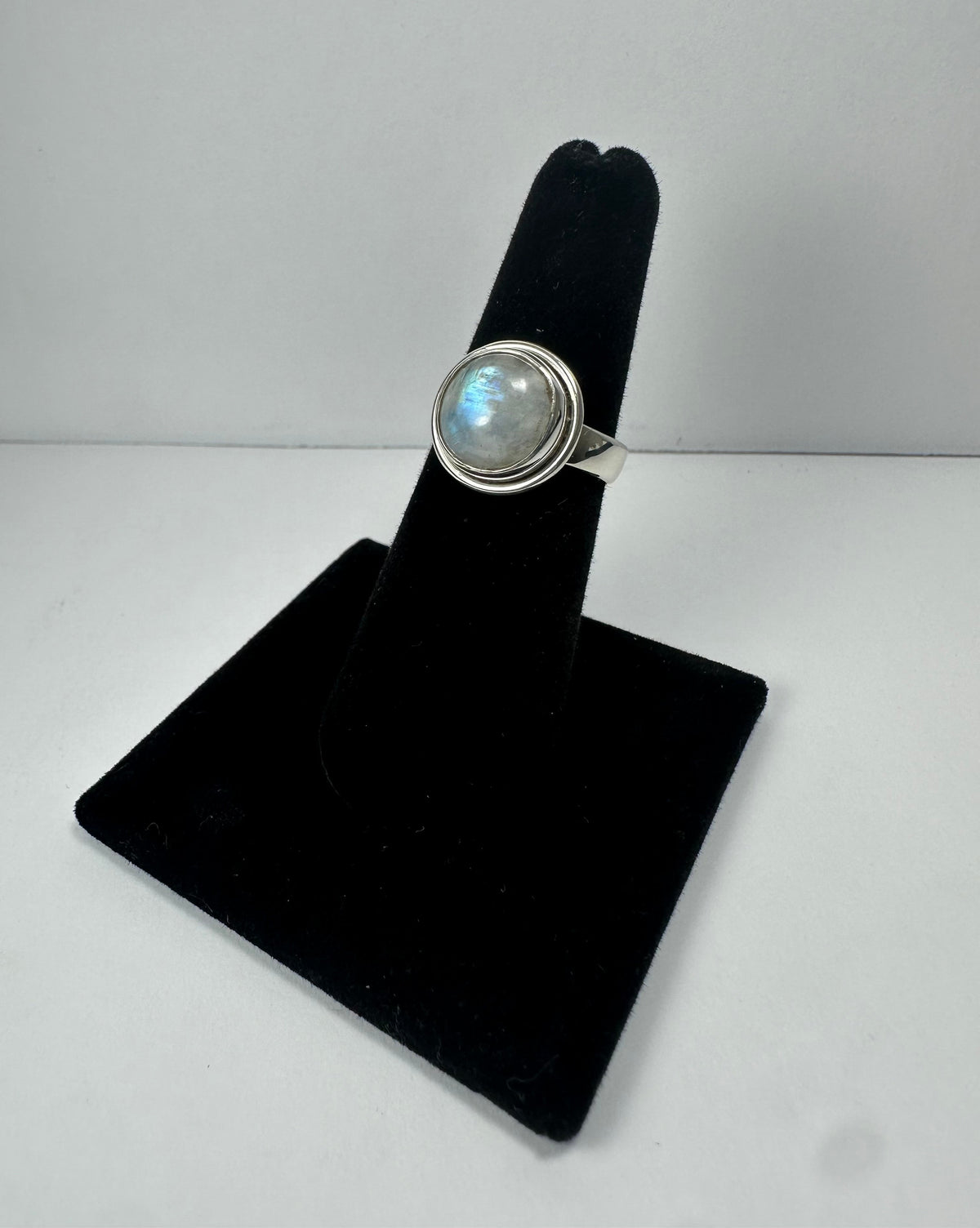 Silver ring with a round iridescent moonstone set in a smooth bezel, displayed upright on a black velvet stand against a plain white background.