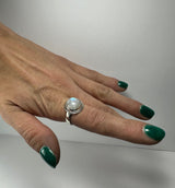 Hand with dark green nail polish wearing a silver ring featuring a round, polished moonstone cabochon with subtle blue and white hues.