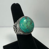 Silver ring with an ornate filigree band featuring a large, rounded turquoise stone with natural veining, displayed on a black velvet finger stand.