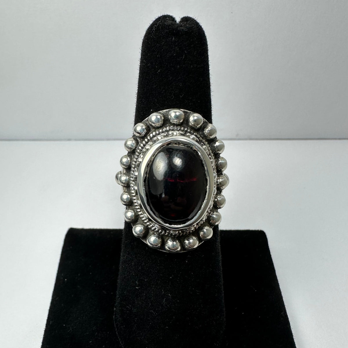 Silver ring with a polished oval dark red garnet center stone, surrounded by a beaded silver border, displayed on a black velvet stand.