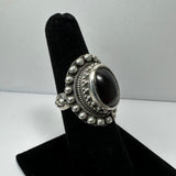 Silver ring with an oval dark red garnet gemstone centerpiece, surrounded by decorative silver bead and twisted rope detailing on a black velvet stand.