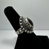 Silver ring with a deep red garnet cabochon center, surrounded by intricate beaded and rope-like ornamental details, displayed on a black velvet stand.