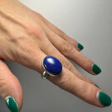Hand with dark green nail polish wearing a silver ring featuring an oval polished deep blue lapis lazuli stone with subtle gold flecks