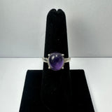 Silver ring with a polished oval amethyst gemstone set in four prongs, displayed upright on a black velvet ring stand against a white background.
