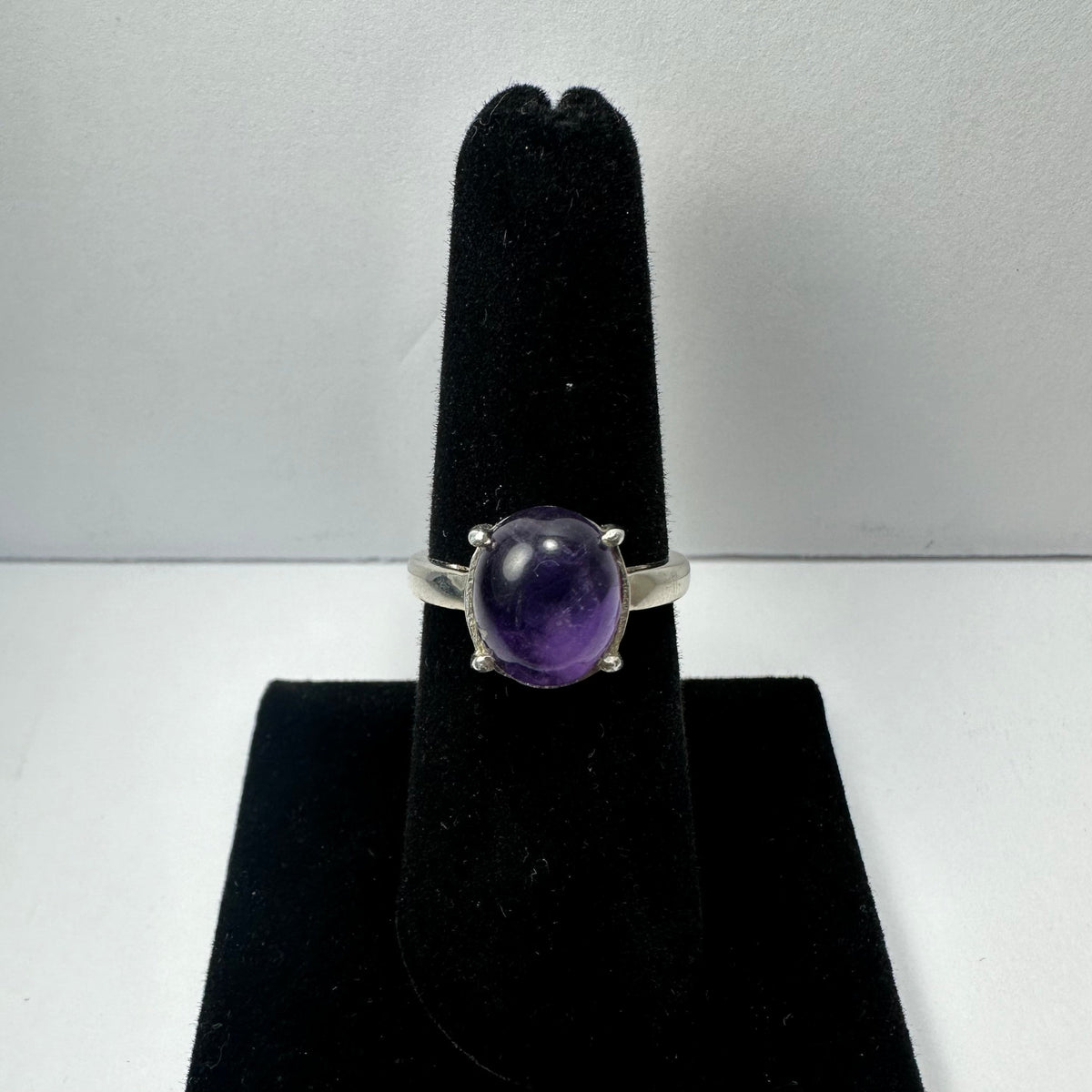 Silver ring with a polished oval amethyst gemstone set in four prongs, displayed upright on a black velvet ring stand against a white background.