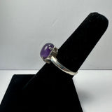 Silver ring with a polished oval purple amethyst gemstone set in a four-prong setting, displayed on a black velvet finger stand