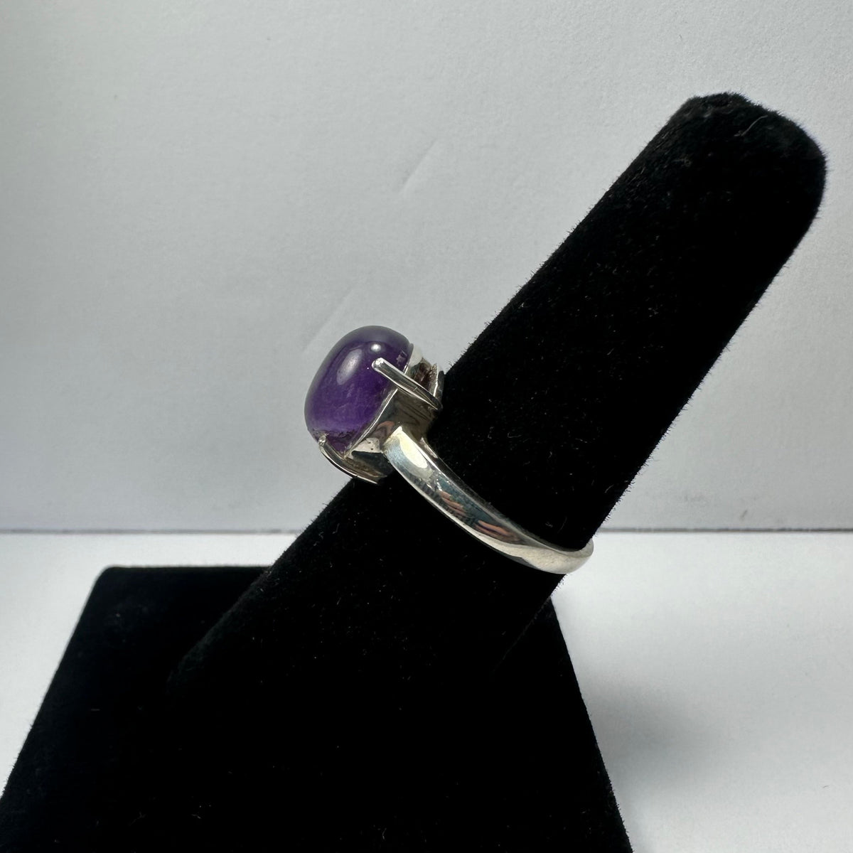 Silver ring with a polished oval purple amethyst gemstone set in a four-prong setting, displayed on a black velvet finger stand