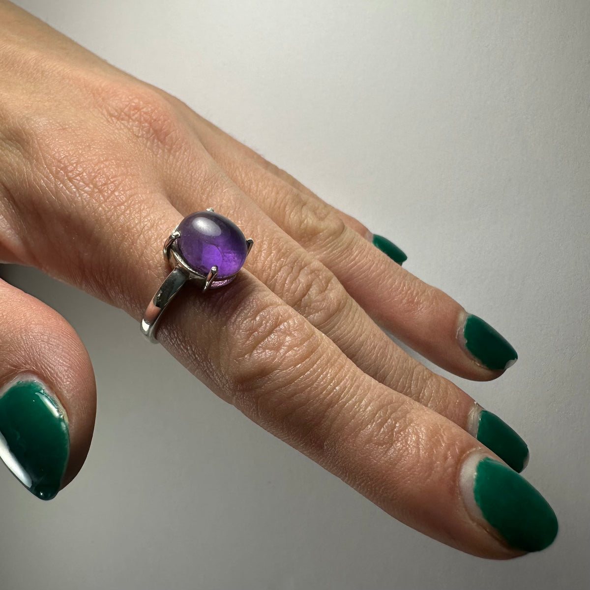 Hand with green nail polish wearing a silver ring featuring a round, polished amethyst stone on the middle finger.