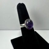 Silver ring with a polished deep purple amethyst oval stone set on a black velvet finger display against a plain gray background.