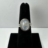 Silver ring featuring an oval moonstone with white and blue-gray marbling, displayed on a black velvet ring holder against a neutral background.