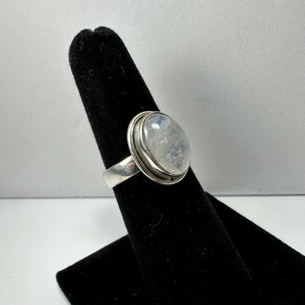Silver ring with a round, translucent moonstone cabochon set in a smooth bezel, displayed on a black velvet ring stand against a plain background.