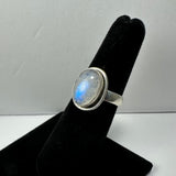 Silver ring with an oval moonstone displaying a blue sheen, mounted on a black velvet ring holder against a plain light background.