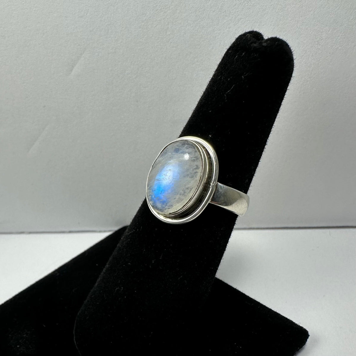 Silver ring with an oval moonstone displaying a blue sheen, mounted on a black velvet ring holder against a plain light background.