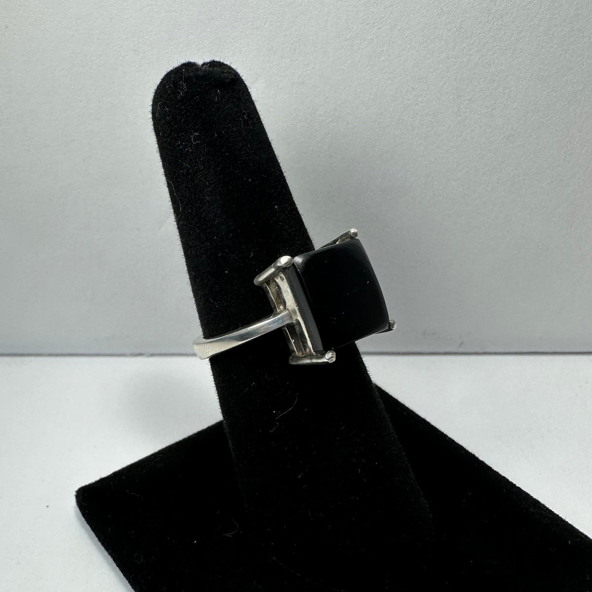 Silver ring with a square black onyx stone set in four prongs, displayed upright on a black velvet ring holder against a plain white background.