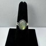 Silver ring with an oval labradorite stone showing green and blue flashes, displayed on a black velvet ring holder against a light background.