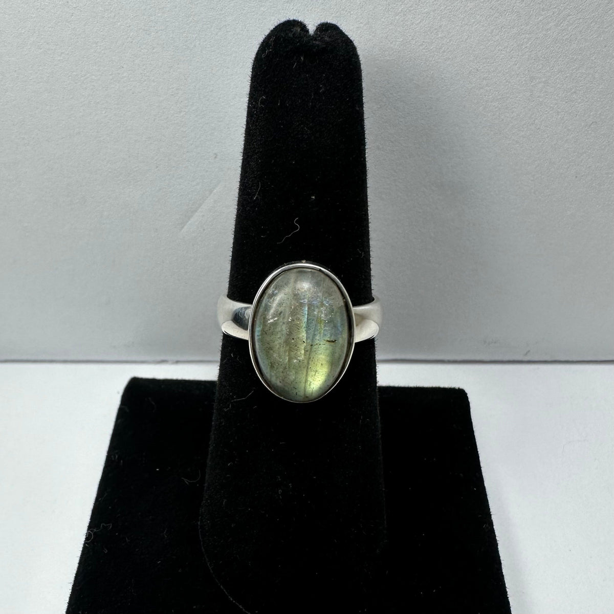 Silver ring with an oval labradorite stone showing green and blue flashes, displayed on a black velvet ring holder against a light background.