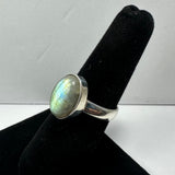 Silver ring with an oval labradorite gemstone displaying blue and green iridescence, showcased on a black velvet finger stand against a white background.