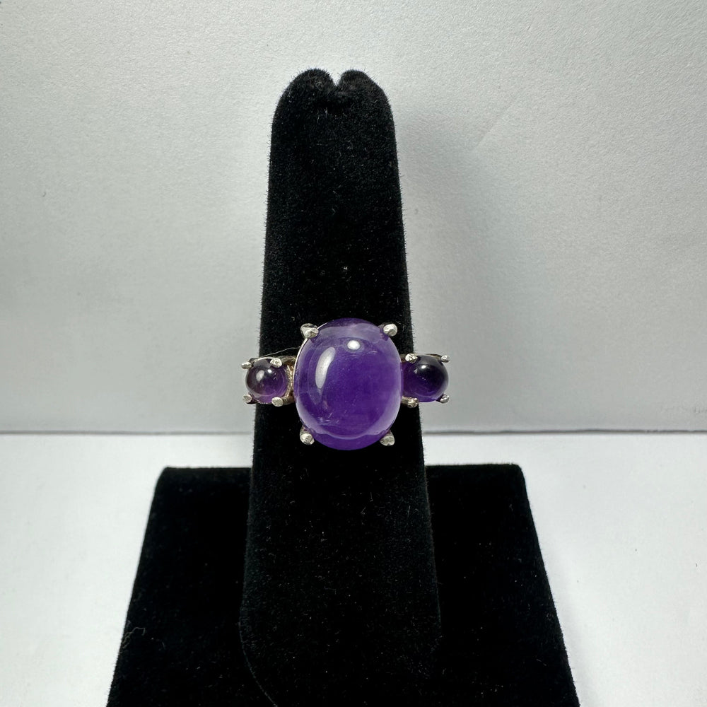 Silver ring with a central oval cabochon amethyst flanked by two smaller round amethyst stones, displayed on a black velvet ring stand against a gray background.