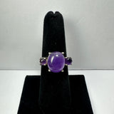 Silver ring with a central oval cabochon amethyst flanked by two smaller round amethyst stones, displayed on a black velvet ring stand against a gray background.