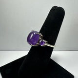 Silver ring with a large smooth oval amethyst stone and a smaller round amethyst set next to it, displayed on a black velvet ring holder against a gray background.