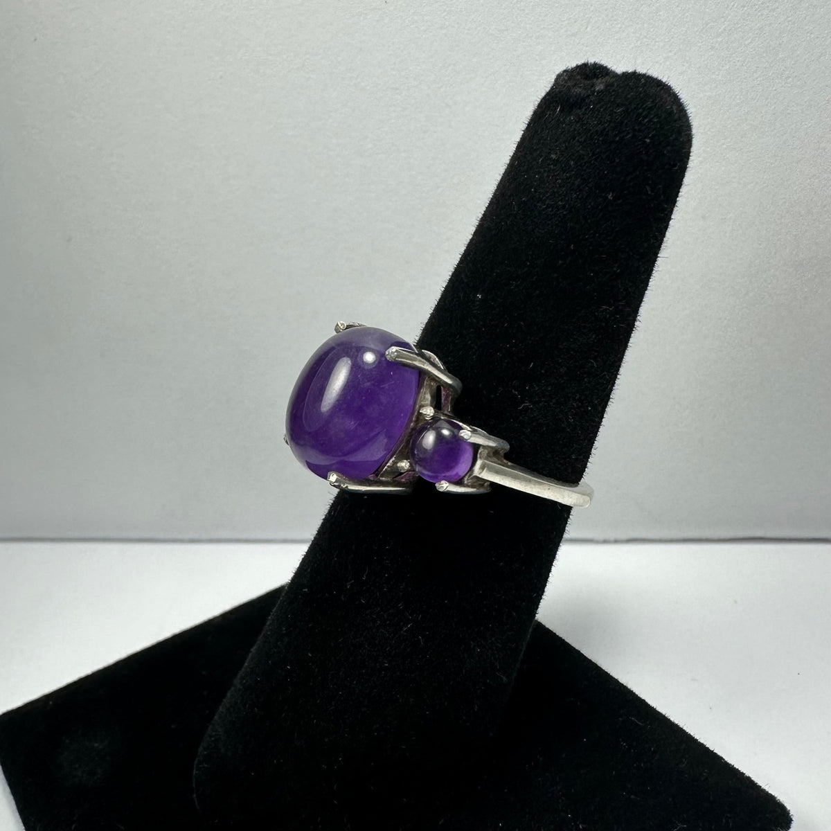 Silver ring with a large smooth oval amethyst stone and a smaller round amethyst set next to it, displayed on a black velvet ring holder against a gray background.