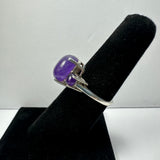 Silver ring with a polished oval amethyst cabochon set in claw prongs, displayed on a black velvet finger stand against a plain light background.