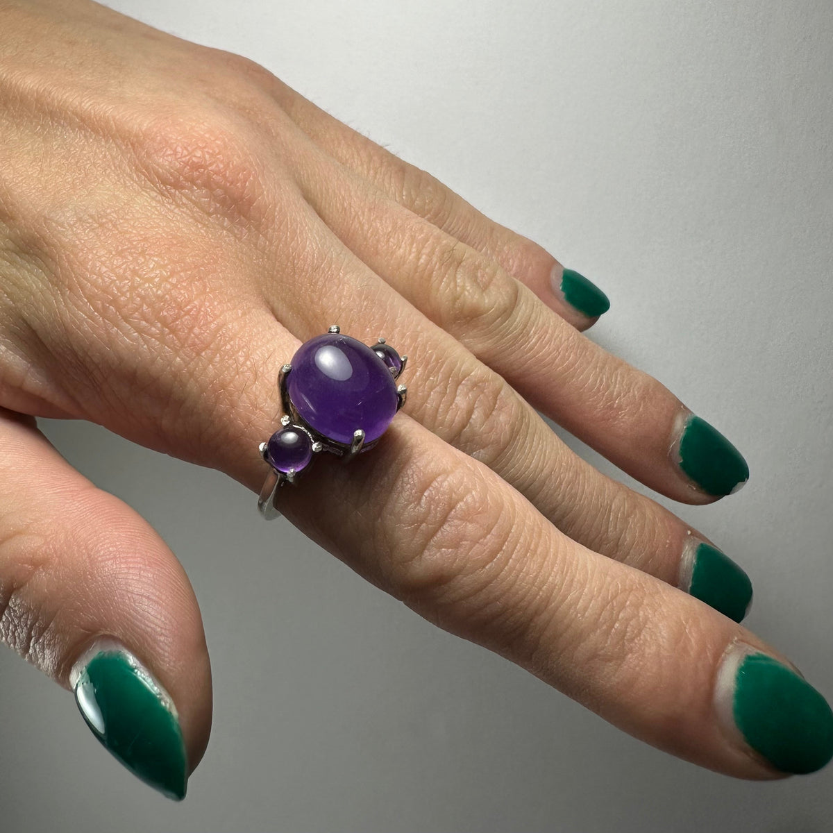 Silver ring featuring a large oval cabochon amethyst center stone flanked by two smaller round cabochon amethyst stones, worn on a hand with green nail polish.