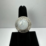 Round moonstone set in an ornate silver filigree ring, displayed upright on a black velvet ring stand against a plain light background.