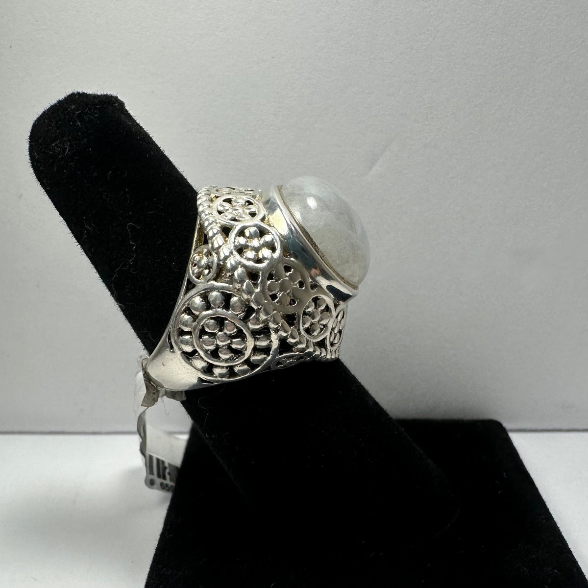 Silver ring with intricate floral filigree details and a smooth, rounded moonstone cabochon set on top, displayed on a black velvet finger stand.