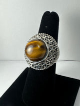 Silver ring with an ornate filigree band featuring a large, round tiger's eye stone with golden-brown and amber hues displayed on a black velvet finger stand.