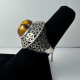 Silver ring with ornate floral filigree details and a polished tiger's eye cabochon stone, displayed on a black ring stand against a plain background.