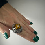 Close-up of hand wearing a size 6 ring with a polished round tiger's eye stone set in an intricate silver floral-patterned band, against a plain background.