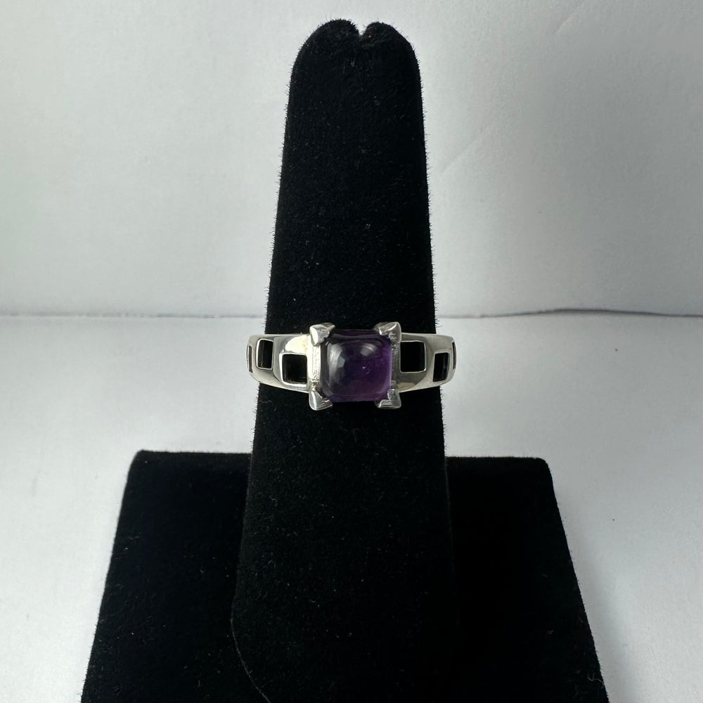 Silver ring with a square amethyst centerpiece set in prongs and a band featuring a geometric cutout pattern, displayed on a black velvet stand against a white background.