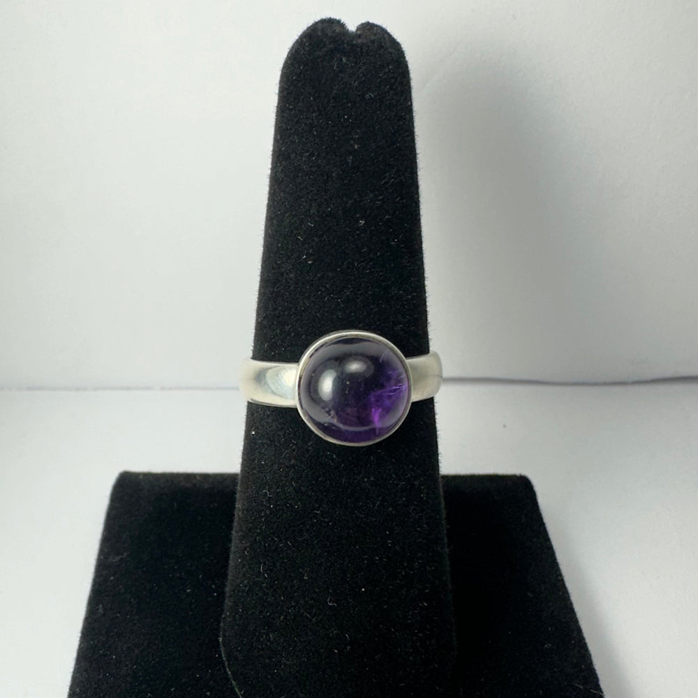 Silver ring with a polished round amethyst cabochon set in the center, displayed on a black velvet ring holder against a white background.