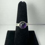 Silver ring with a polished round amethyst cabochon set in the center, displayed on a black velvet ring holder against a white background.