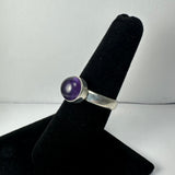 Silver ring with a polished round purple amethyst cabochon set on a simple band, displayed on a black velvet finger stand against a white background.