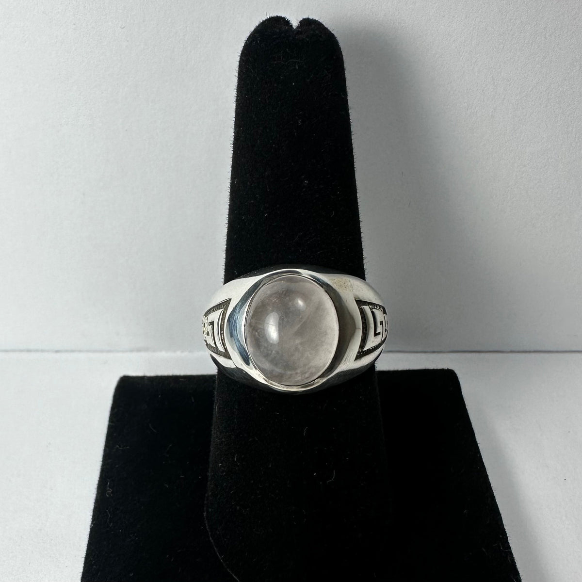 Silver ring with a smooth, translucent, round moonstone set in the center and geometric patterns engraved on the wide band sides, displayed on a black velvet ring stand.