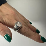 Silver ring with a smooth round moonstone set in a detailed geometric band worn on the middle finger with dark green painted nails.